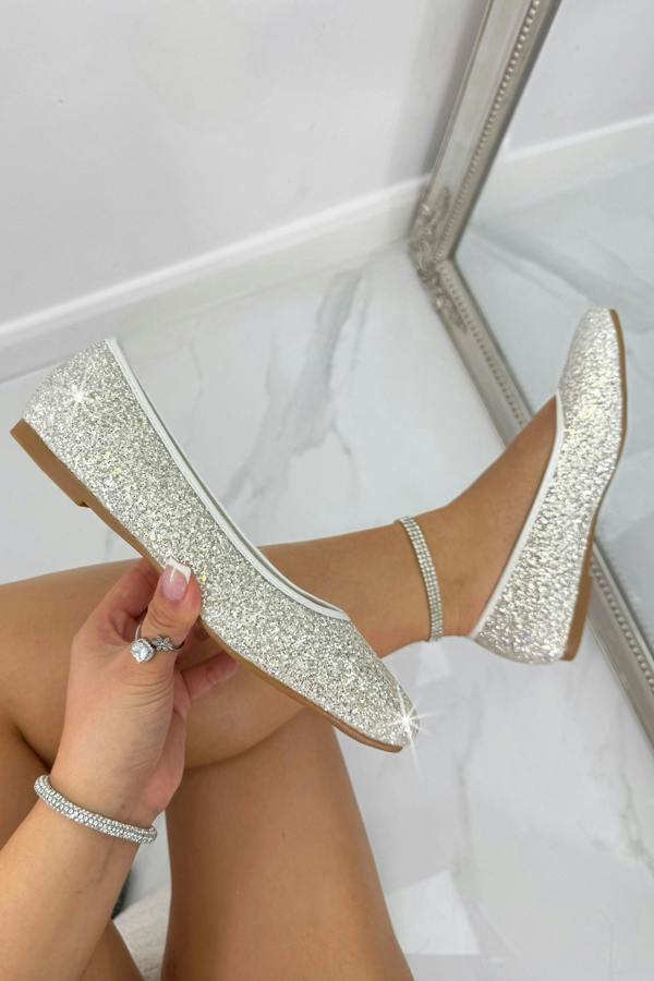Sparkly white shoes being held by a hand with a ring, on a white background.