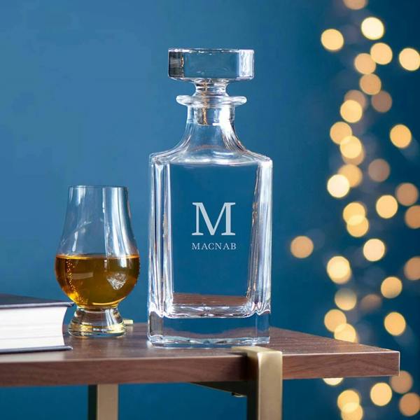 A heavy-weight square glass decanter with a round stopper sits on a wooden table next to a short glass of amber whiskey and a book, featuring a bold monogram 'M' and the name 'MACNAB' engraved on the front against a blue background with festive bokeh lights.