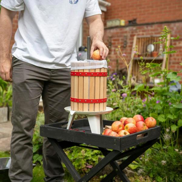 KuKoo fruit crusher with wooden barrel design, red accents, and a person placing an apple into the press, surrounded by fresh apples