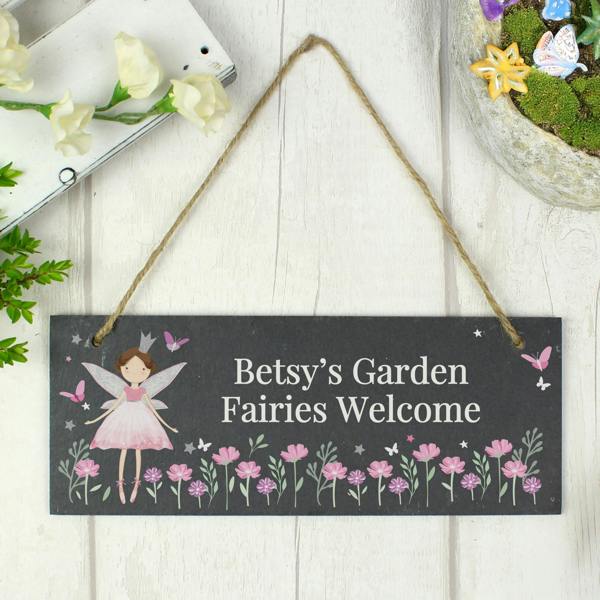 A rectangular natural dark slate hanging sign hangs from a rustic jute twine loop against a white wood panel background. The left features a printed fairy princess illustration next to the custom text 'Betsy's Garden, Fairies Welcome' above a border of pink wildflowers and butterflies. A miniature fairy garden container is partially visible in the top right corner.