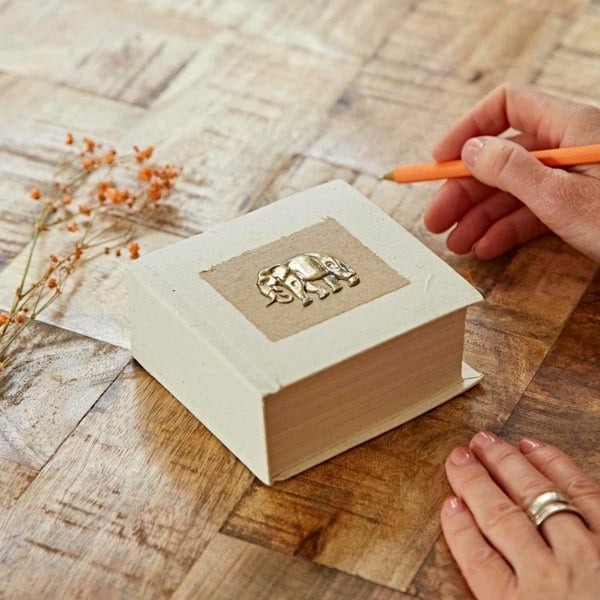 A hand with an orange pen is ready to write in a small, decorative Elephant Dung Memo Block with an embossed elephant on the cover, resting on a wooden table. Nearby are scattered small dried flowers.