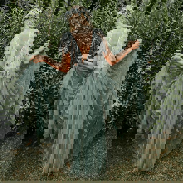 A young girl wearing a sage green Bohemian style dress with a layered tulle skirt and lace details, standing in front of a bush.