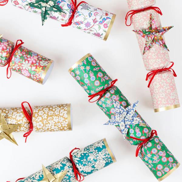 A selection of luxury Liberty print Christmas crackers tied with red velvet bows and decorated with origami stars scattered on a clean white background.