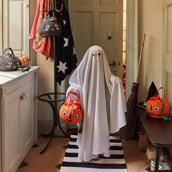 A child dressed in a white ghost costume holding a vibrant orange woven pumpkin trick or treat bag in a beautifully styled hallway.
