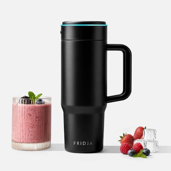 A sleek black Fridja f600 Stainless Steel Portable Blender stands between a glass of pink berry smoothie with berries and mint, and a pile of fresh strawberries, blueberries, raspberries, and ice cubes on a light background.