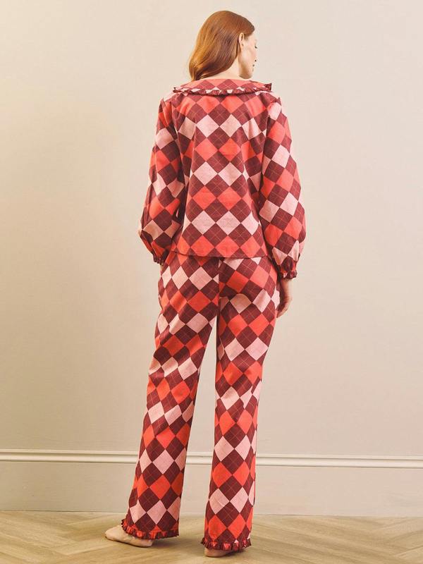 A woman stands with her back to the camera wearing a matching long-sleeve pyjama set with a red, pink, and burgundy harlequin diamond pattern, set against a neutral beige wall and light wood floor.
