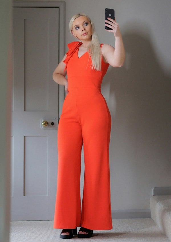 This is a young lady wearing a Flame red coloured V neck jumpsuit, with wide legs and a bow on one shoulder. She is wearing black open-toed sandals and is taking a selfie on a black phone.