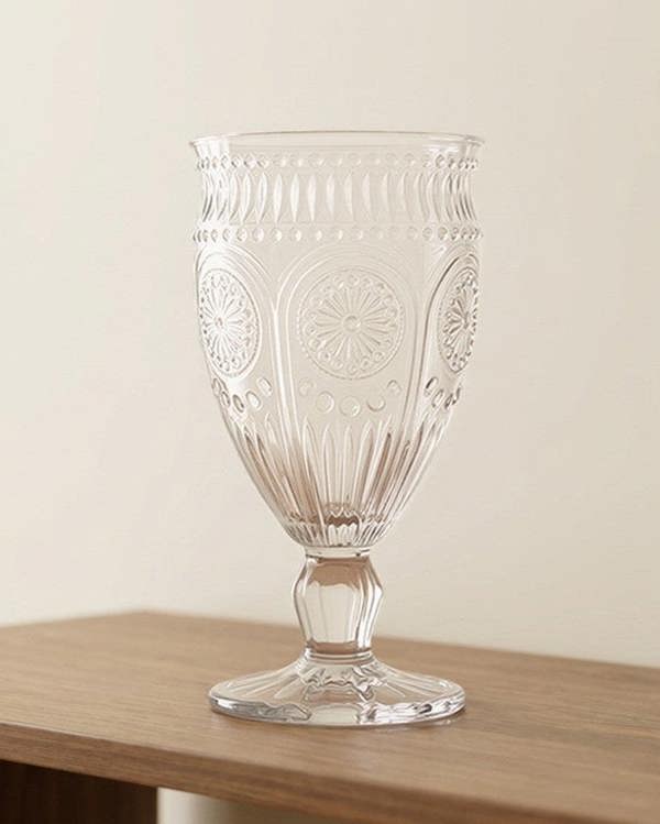 A clear glass goblet with etched floral patterns sits on a light brown wooden shelf against a warm beige wall.