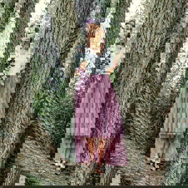 Young girl in a white top and mauve skirt standing between two tree trunks in a forest.