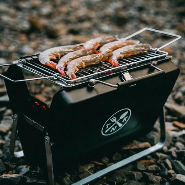 Portable Notebook BBQ with fresh king prawns grilling on a rocky beach.