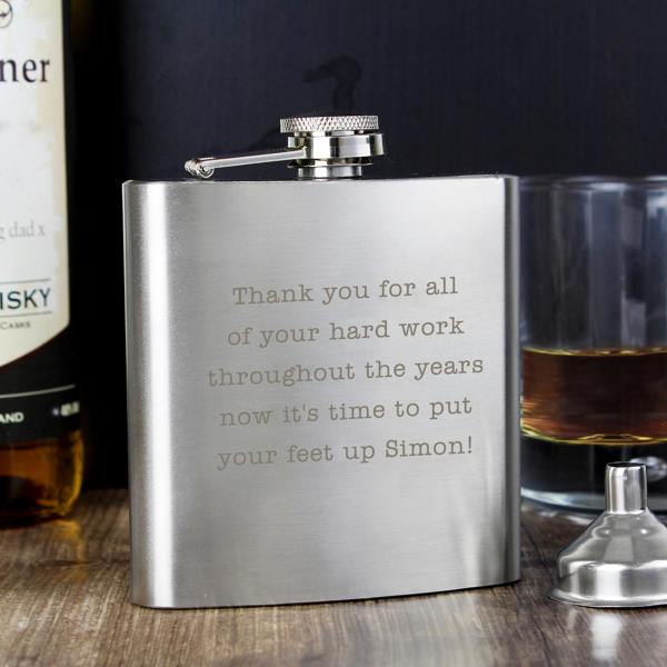 A personalised stainless steel hip flask engraved with a retirement message next to a tumbler of whiskey and a funnel.