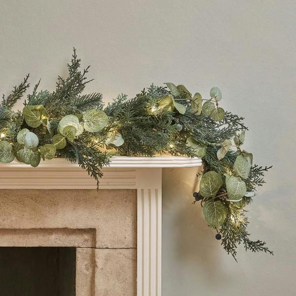 An oversized pre-lit Christmas garland with eucalyptus, pine, and warm white LED lights arranged across a classic stone fireplace mantel.