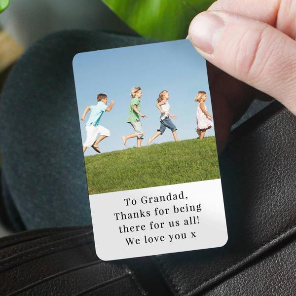 A hand sliding a metal photo wallet card into a brown leather wallet, printed with a photo of kids running and text reading 'To Grandad...'.