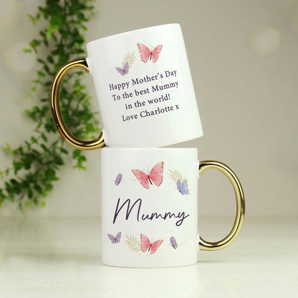 Two custom white ceramic mugs with shiny gold handles stacked together, showing a butterfly pattern, the front text Mummy, and a Mother's Day message on the reverse.