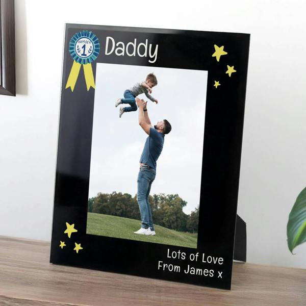 A sleek black 5x7 portrait photo frame featuring a blue 'Number 1' rosette icon and yellow stars. Inside is a photo of a father lifting his son into the air. The bottom right is personalised with the text 'Lots of Love From James x'.