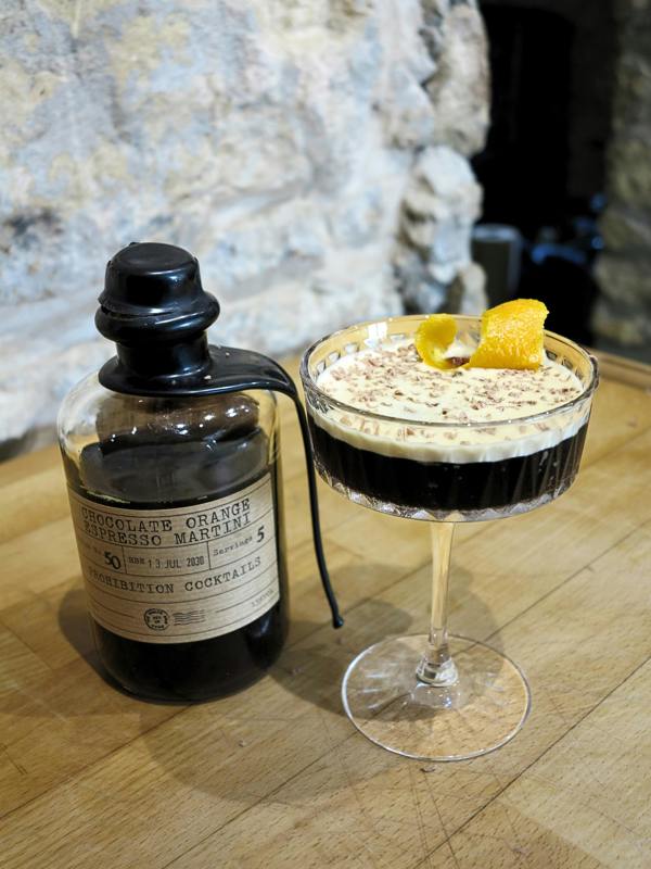 A bottle of chocolate orange espresso martini mixed liquor by Prohibition Cocktails next to a prepared cocktail in a coupe glass, garnished with a citrus peel, on a wooden table with a rustic stone wall background.