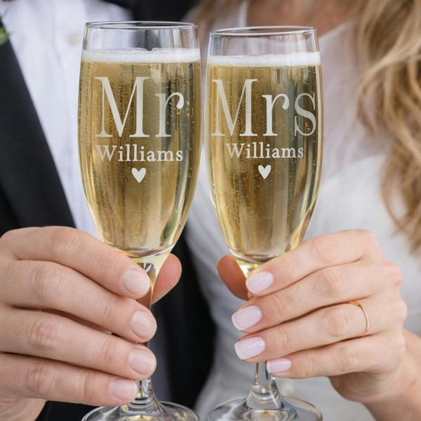 A close-up shot of a bride and groom holding up a matching pair of personalised champagne flutes by Abode Home & Gifts, engraved with "Mr Williams" and "Mrs Williams" above a small heart, filled with bubbling champagne.