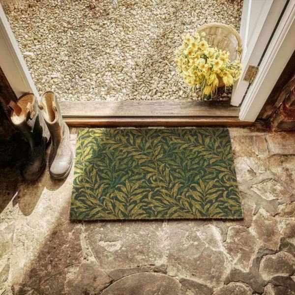 Doormat with leaf pattern on stone floor, shoes, and basket with flowers.