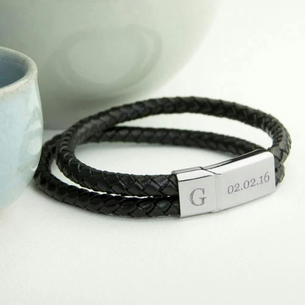 A close-up of a personalised men's dual woven leather bracelet in black with a polished silver metal clasp, showing fine custom-engraved text on its surface.