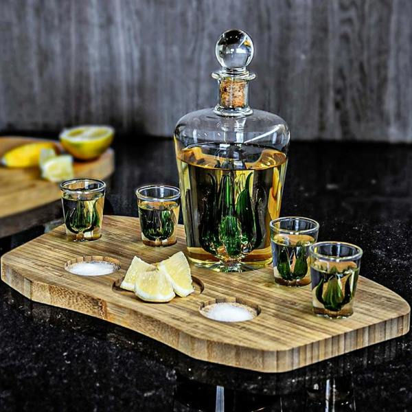 A luxury glass tequila decanter filled with golden liquid and 4 matching shot glasses, featuring green glass agave plants inside, displayed on a bamboo serving board with lemon slices and salt.