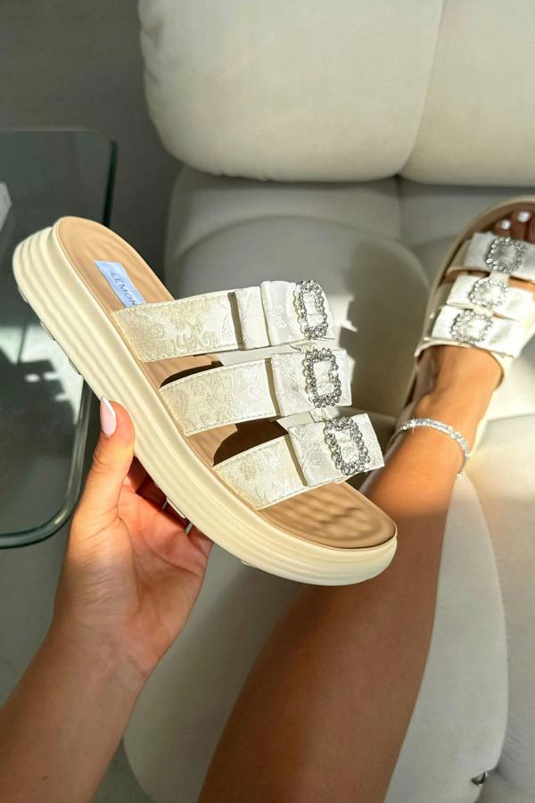 Beige glittery sandals with rhinestone straps held by a hand, on a neutral background.