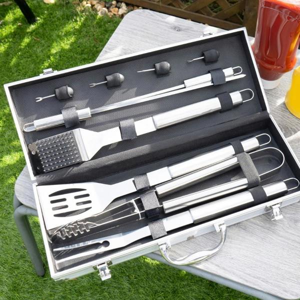 An open aluminium brief-style carry case displaying a clean 10-piece stainless steel barbecue tool set with a spatula, tongs, fork, skewers, and cleaning brush on a grey outdoor table.