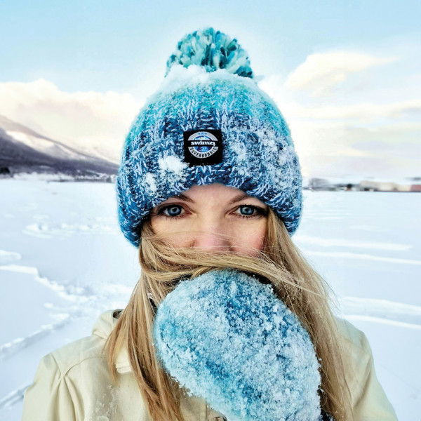 Woman stands amongst a snowy landscape, wearing a striped bobble hat in a  shades of dark to light turquoise blue with a large pom-pom. The hat also has a deep ribbed headband and contains reflective yarns and a luxury sherpa fleece lining.