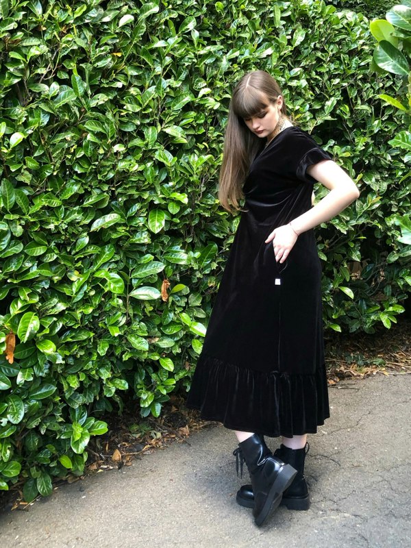 This is a black frill velvet dress worn by a model with dark long straight hair against a Japonica hedge. It has a knot and plunge v neck with short sleeves. The model is facing the left side and she is looking down at her hand in her pocket. She is wearing black boots.