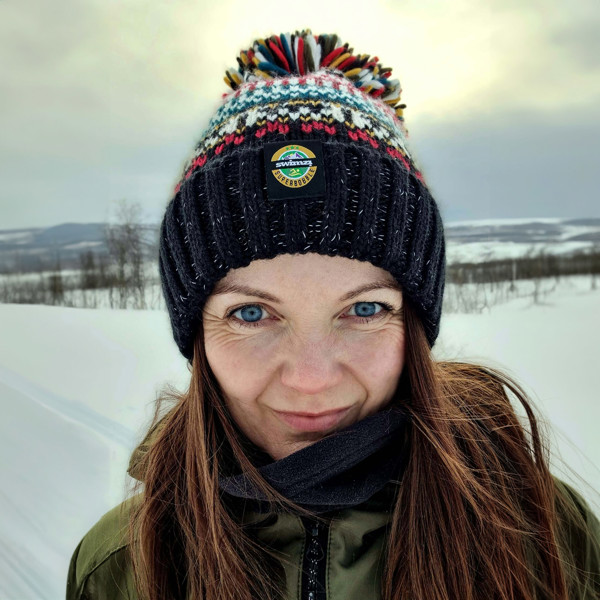 Woman in a wintery landscape backlit by a fading sunset is wearing a traditional Nordic design chunky knit Super Bobble Hat. The black base knit is accented by dark red, deep teal and warm cream.