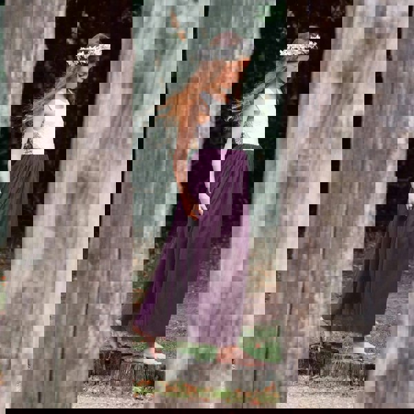 Young girl in a white top and purple skirt standing between two wooden posts in a natural setting.