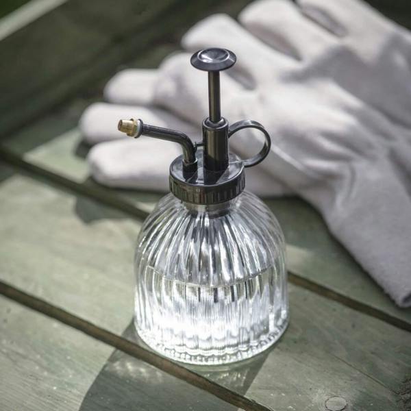 Ribbed clear glass indoor plant mister with black pump mechanism sitting on a wooden bench beside gardening gloves.