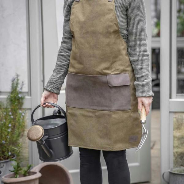 Abode Home & Gifts khaki canvas garden apron with brown front tool pockets, worn whilst holding a black watering can and hand fork.