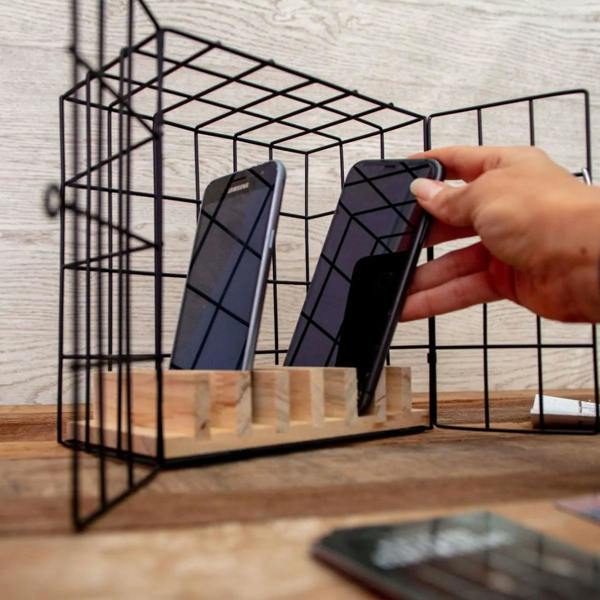 A close-up shot of the Phone Escape Room Game, featuring a black wire cage on a wooden base. A hand is shown sliding a smartphone into one of the wooden slots inside the cage. The background is a soft, out-of-focus living room setting.