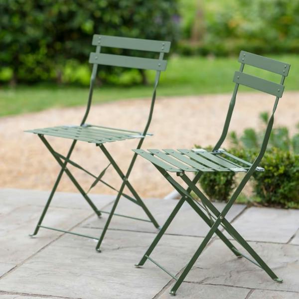 A pair of thistle green metal folding bistro chairs positioned on a light stone patio in a lush garden.