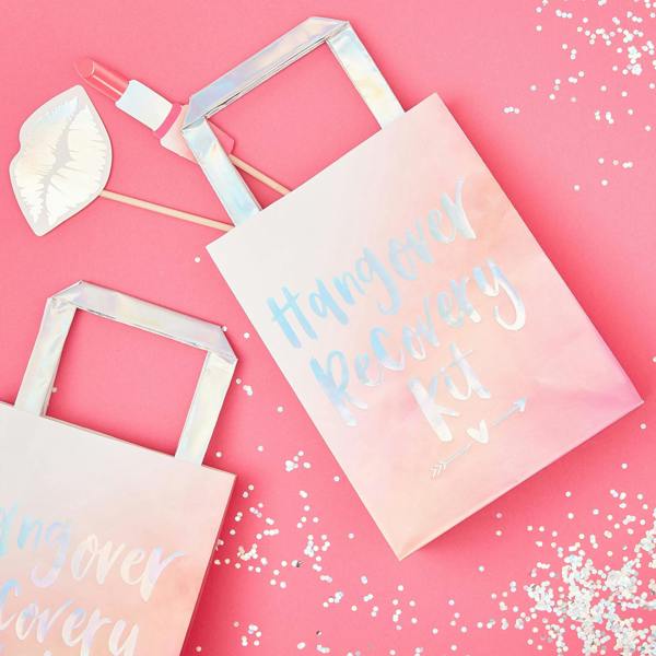Flat lay view of two pink ombré Hen Party Hangover Recovery Kit bags with iridescent foil script and handles alongside a photo booth lip prop and silver confetti scatter.