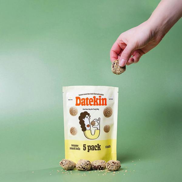 Hand holding a Datekin sesame snack ball above a package of Datekin snack balls on a green background