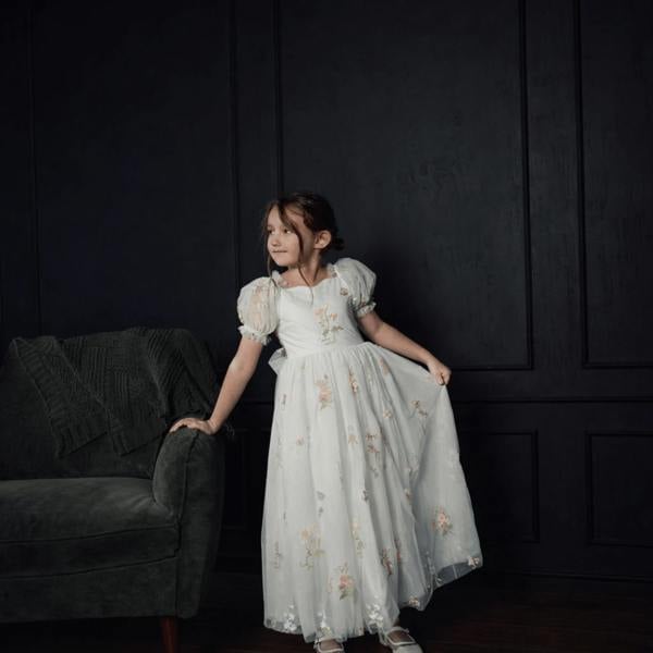 Young girl in a floral dress standing against a dark wall with a gray couch.