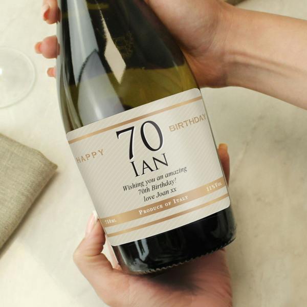 A close-up of a person's hands holding a 75cl bottle of Italian Prosecco with a personalised cream label reading 'Happy 70th Birthday Ian' with gold details.