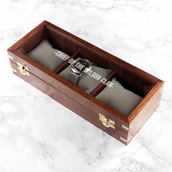 An overhead view of a rich brown wooden watch box with a glass lid displaying three soft grey cushions and one silver watch, featuring custom white text etched on the glass.