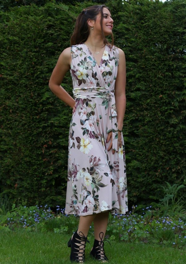 This is a young lady wearing a floral printed dress in pale pink. It has a mock V neck style top, which has gathers under the bust and the shoulders with a tie on the front. The model is wearing black lace up heels, she has her hand on her hip and is looking into the distance, silhouetted against a lush garden.