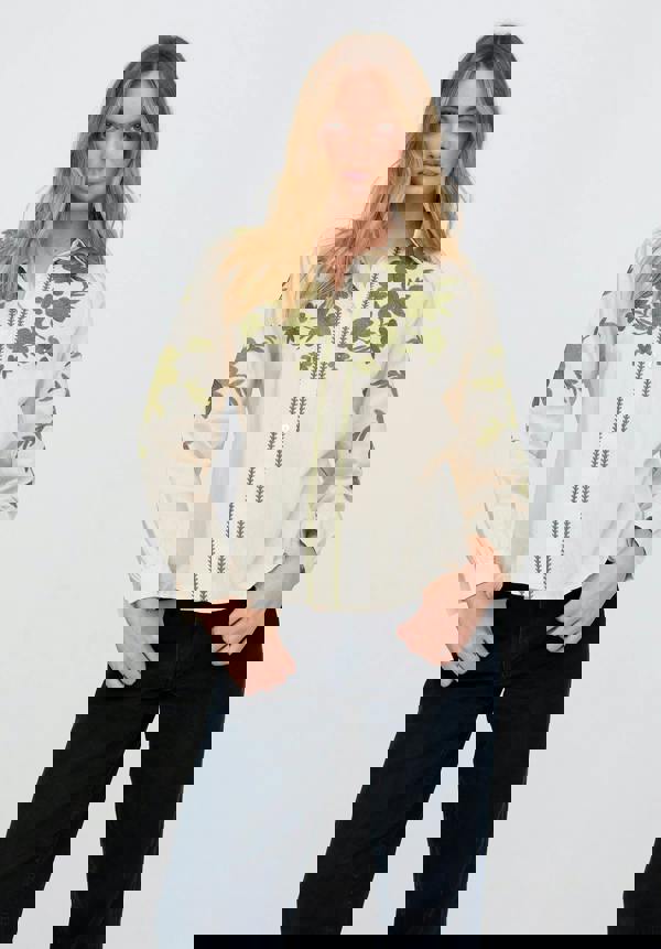 Woman wearing a cream blouse with green floral embroidery on a plain background