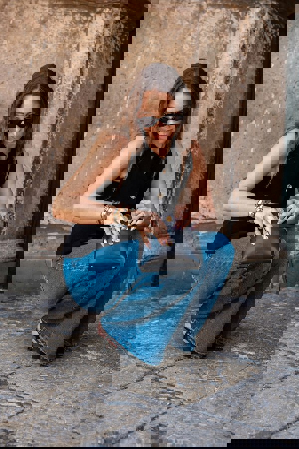 Woman in black sleeveless top and blue jeans holding a black clutch against a stone wall.