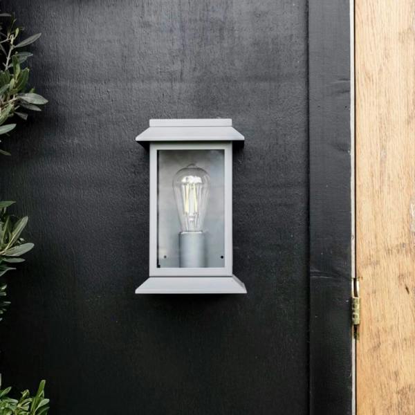 Modern charcoal steel lantern wall light with a matte finish and clear glass frame, mounted on a black wooden exterior wall next to a light oak door.