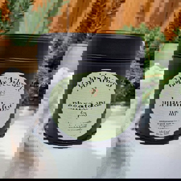 Candle tin labeled 'Ralph's Orchard Enchanted Forest Pine' on a light surface with plants in the background.