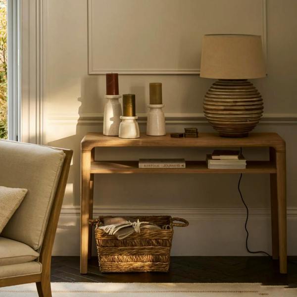 A styled curved oak console table featuring a lower shelf, decorated with a large ceramic table lamp, candles, and a woven storage basket underneath.