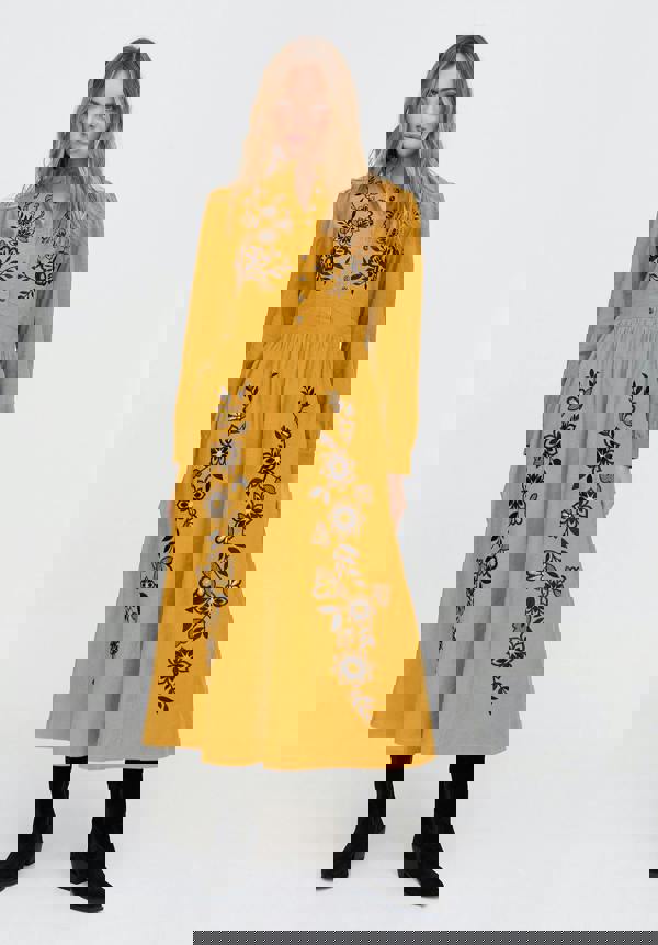 Woman wearing a mustard yellow dress with black floral embroidery on a white background