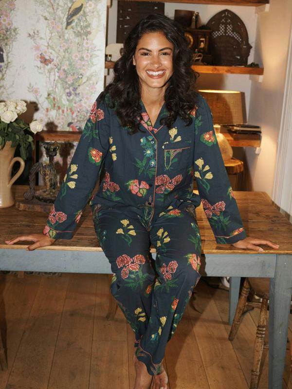 A woman wearing a dark teal floral pyjama set with pink, red, and green flowers, sitting on a rustic wooden table in a cozy, vintage-style room.