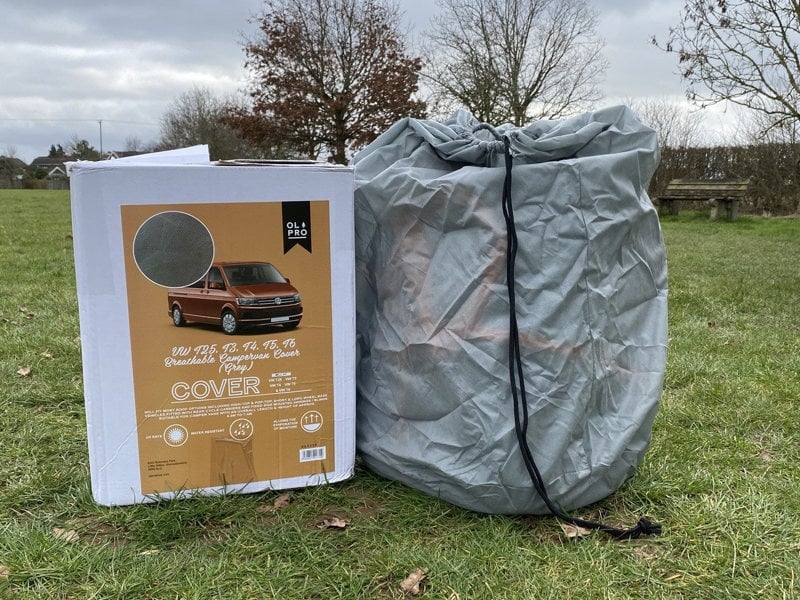 The VW T25/T3/T4/T5/T6 Campervan Cover Grey by OLPRO in its drawstring bag with it's packaging displayed to the side of it in a field.