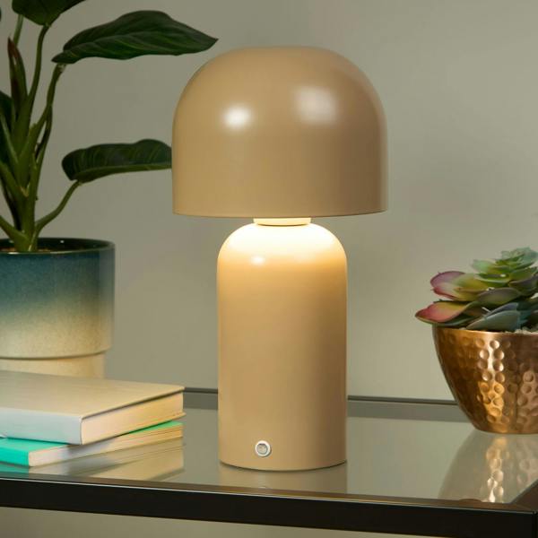 A taupe colored rechargeable touch table lamp with a rounded shade is switched on, providing warm white light. It is placed on a reflective surface, possibly a bedside table, with a plant and a bowl in the background.