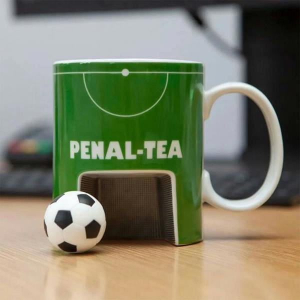 A green ceramic mug designed to look like a football pitch with the text 'PENAL-TEA' printed on it, featuring a built-in cutout goal at the bottom with a mini football next to it on a wooden desk.